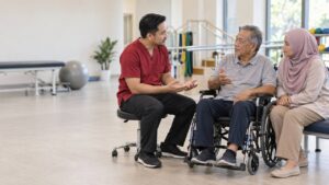 Profesional kesihatan berbincang dengan seorang lelaki berusia yang menggunakan kerusi roda dan seorang peneman di bilik rehabilitasi.